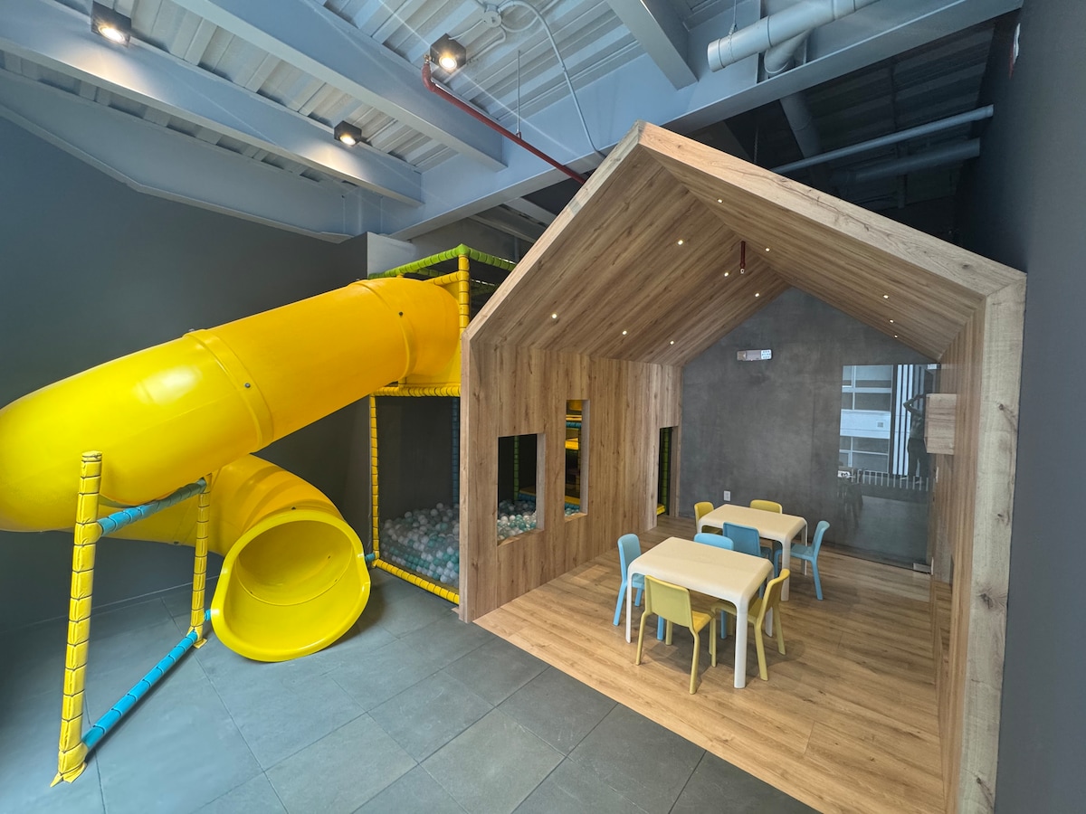 A vibrant play area features a yellow slide leading into a ball pit. A wooden playhouse with large windows is situated nearby, containing a small table and chairs in blue and yellow. The flooring is dark and contrasts with the bright colors of the play equipment.