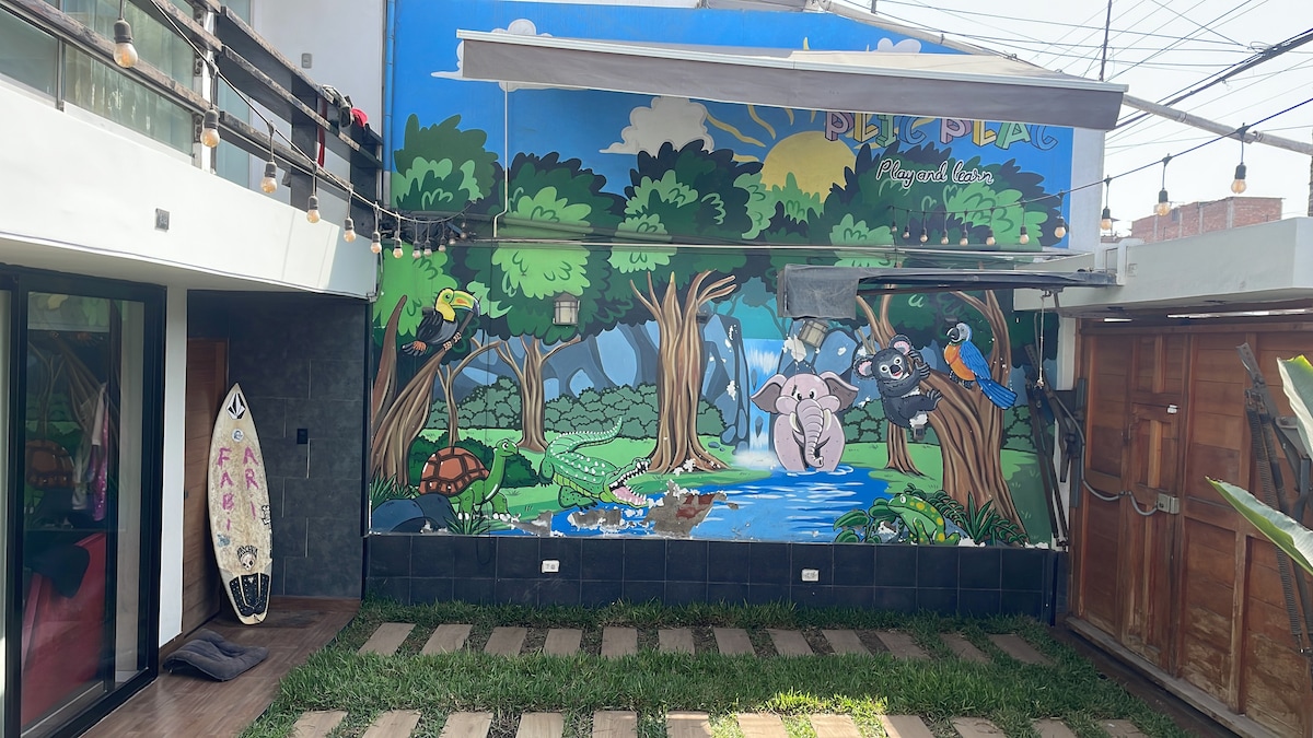 A vibrant mural depicting a serene nature scene is displayed on the wall, featuring animals near a tranquil waterway. The surrounding area includes green grass and a surfboard leaning against the wall, with string lights overhead creating a festive atmosphere.