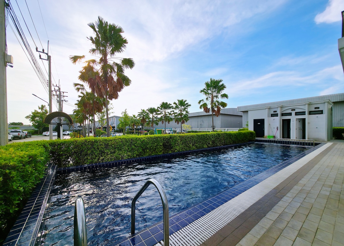 A landscaped pool area is featured with clear blue water reflecting the sky. Surrounding palm trees provide greenery, while the deck is lined with beige tiles. A handrail leads into the pool, and the background includes modern facilities.