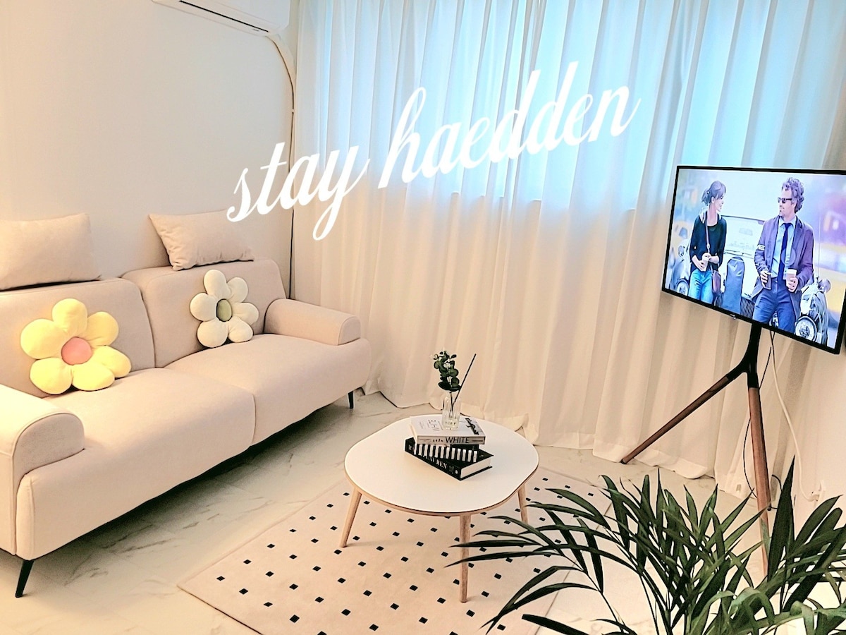 A modern living area is presented, featuring a light-colored sofa adorned with flower-shaped cushions. A round coffee table rests on a patterned rug, while a tall plant adds a touch of greenery. Natural light filters through sheer curtains, enhancing the welcoming ambiance.