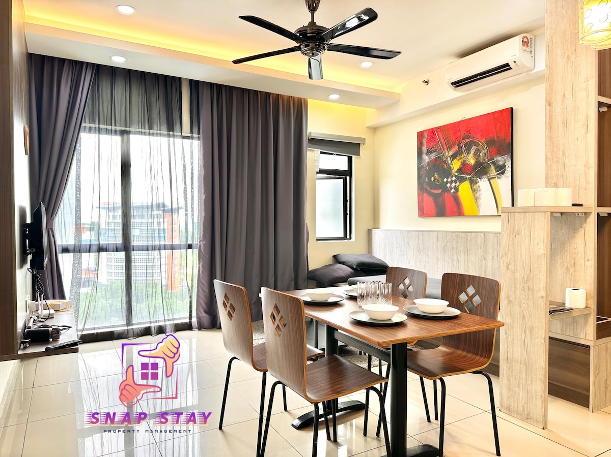 A dining area features a modern table surrounded by four wooden chairs. A large window allows natural light to enter, with light gray curtains partially drawn. A vibrant abstract painting is displayed on the wall, adding a splash of color to the space.
