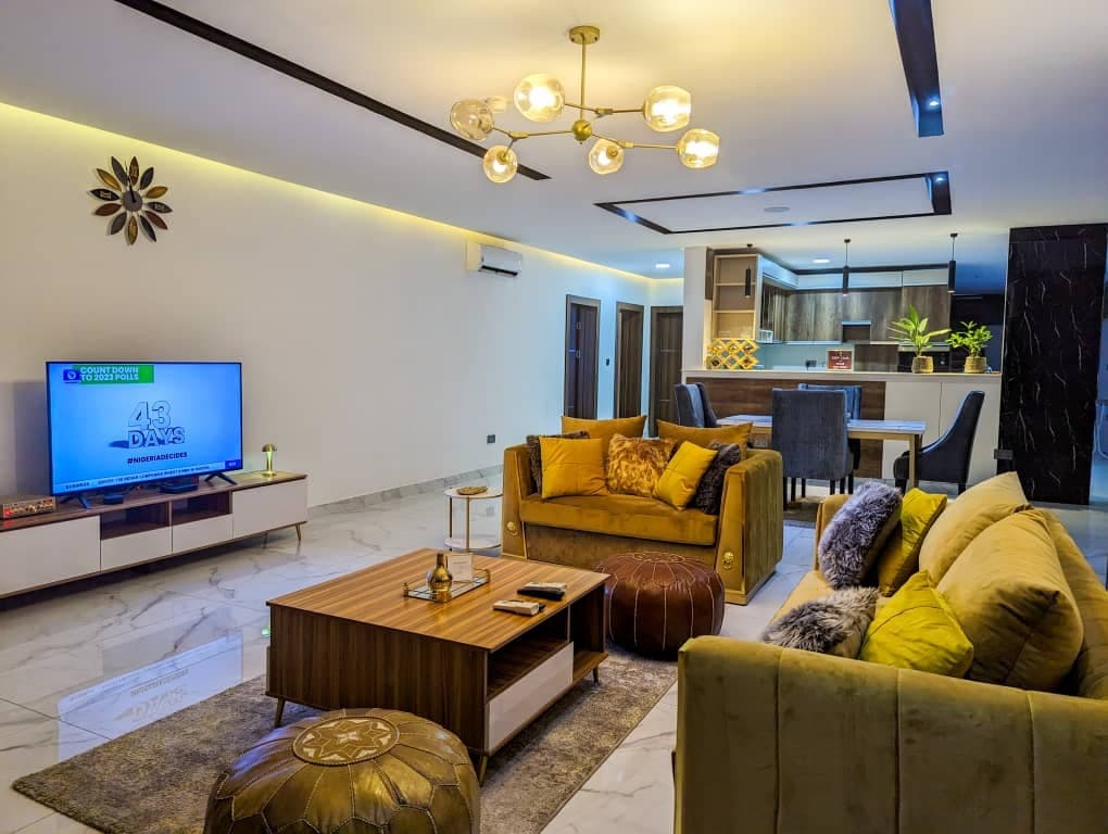 A spacious living area is highlighted by a large television set opposite comfortable seating arrangements with decorative pillows. A coffee table is placed at the center, complemented by stylish poufs. The open layout connects to a dining area visible in the background.