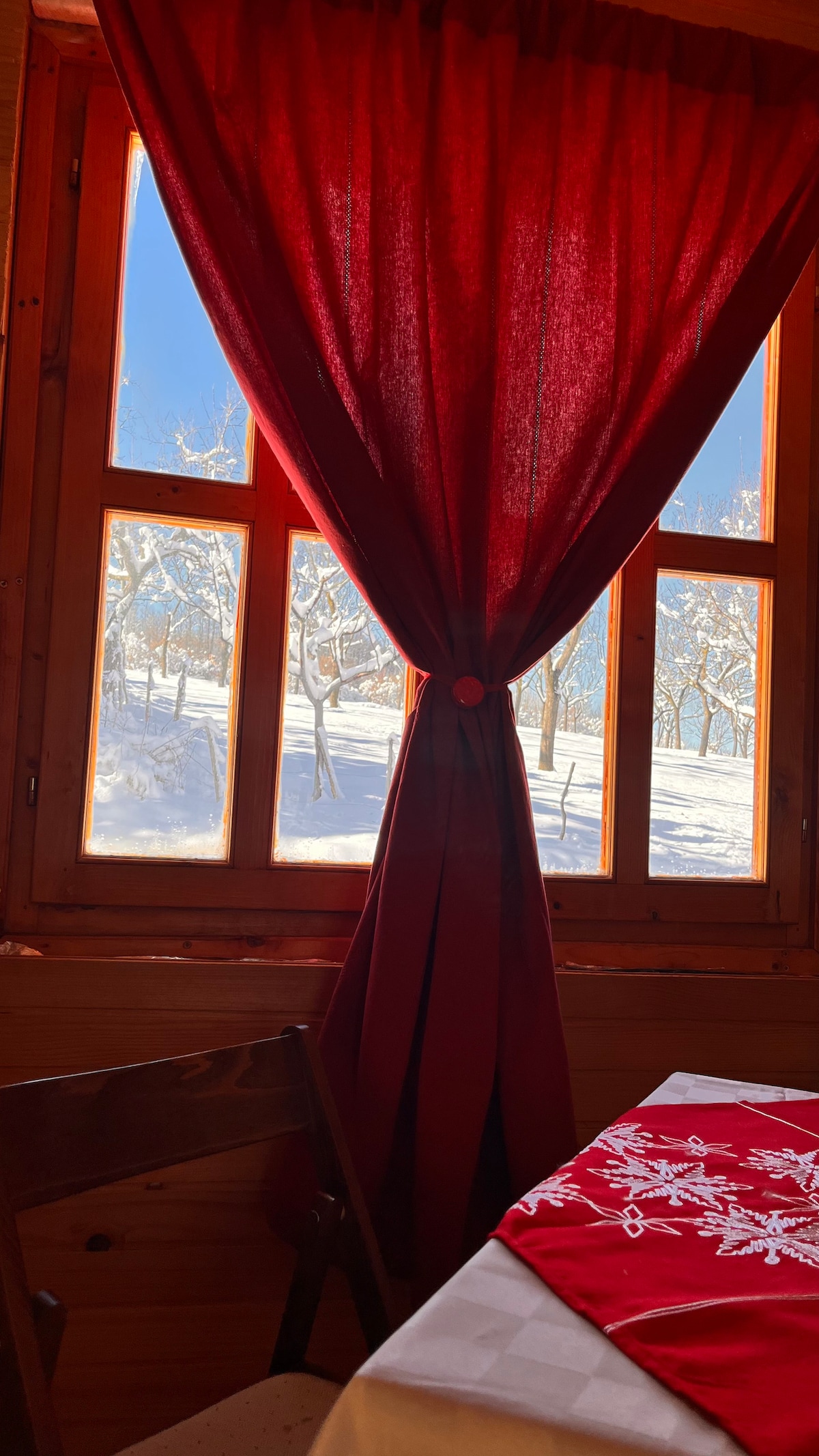A wooden interior features a table covered with a red tablecloth adorned with white floral patterns. Sunlight filters through a window framed in wood, with bright red curtains partially drawn, revealing a snowy landscape outside.