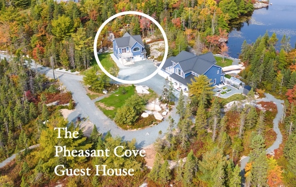 An aerial view of The Pheasant Cove Guest House is presented, showcasing its modern architecture within a landscape of vibrant autumn foliage and a serene lake. A winding path leads to the entrance, emphasizing the tranquil, natural surroundings.