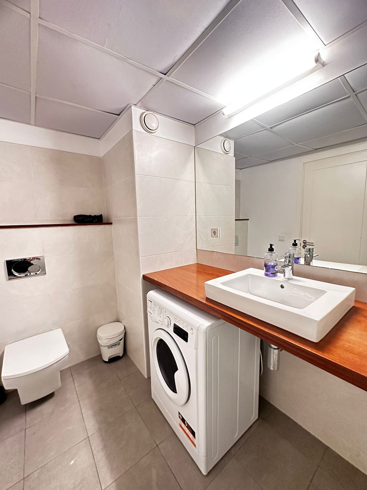 A modern bathroom features light-colored tiled walls and a wooden countertop. A compact washbasin is positioned next to a washing machine. A toilet is situated along the wall, and a large mirror reflects the space's clean lines and functional design.