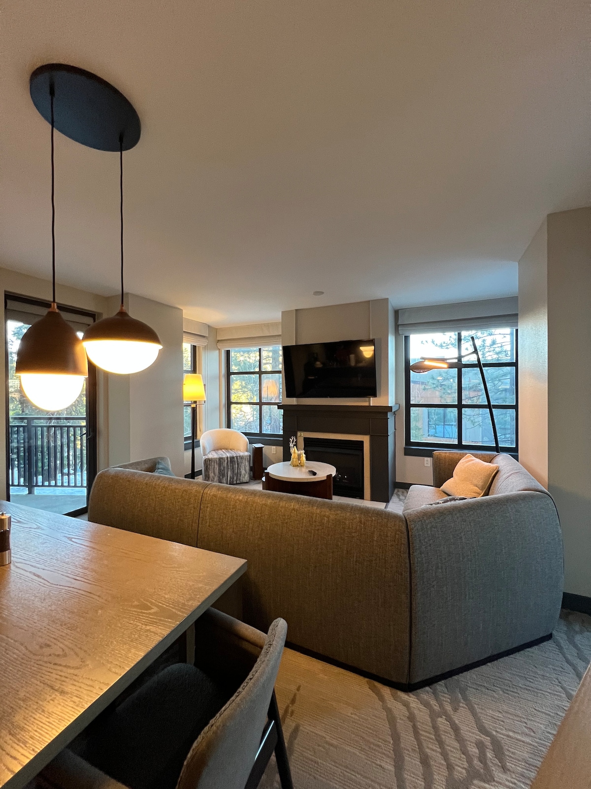 A cozy living area features a soft gray sectional sofa and a contemporary coffee table. A flat-screen television is mounted above a modern fireplace. Large windows allow natural light to fill the space, revealing views of the outdoors. A dining table for six is positioned nearby.