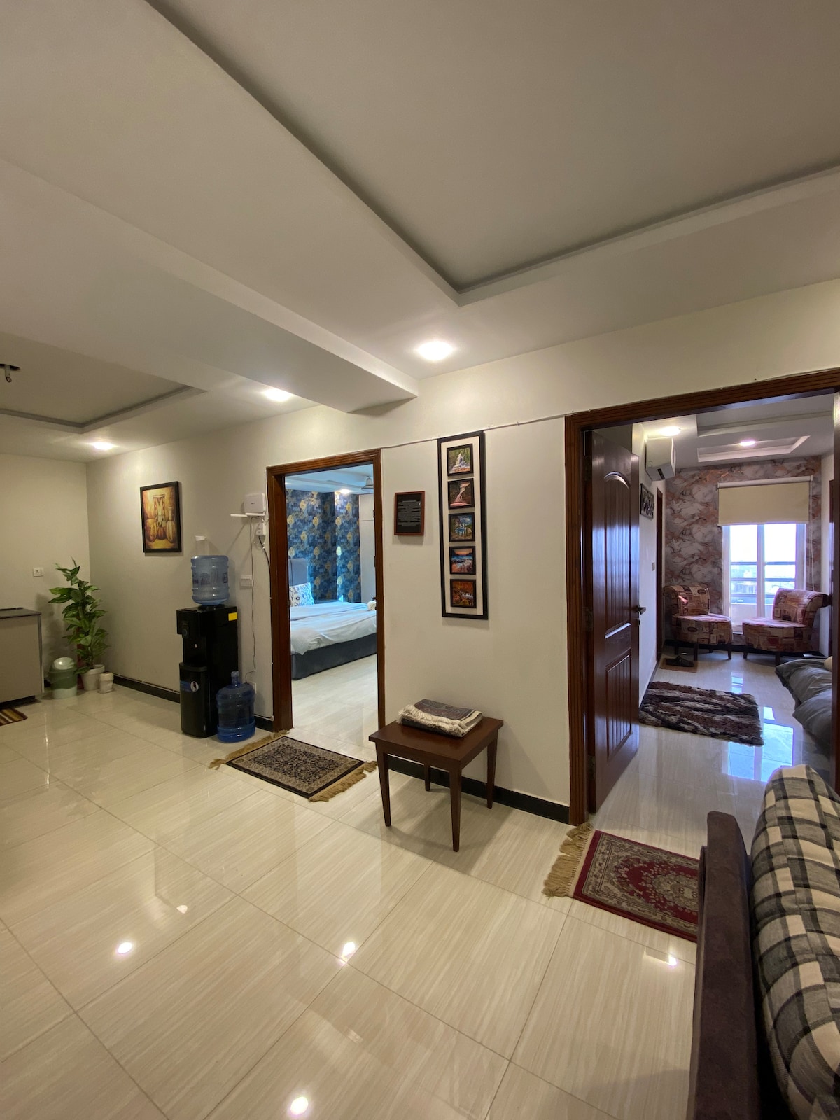 A well-lit entryway connects various living spaces within the apartment. The polished floor reflects the warm lighting, while decorative artwork lines the walls. A small table is positioned beside a water dispenser, leading to a cozy space with a sofa in the foreground and a bedroom visible through an open door.