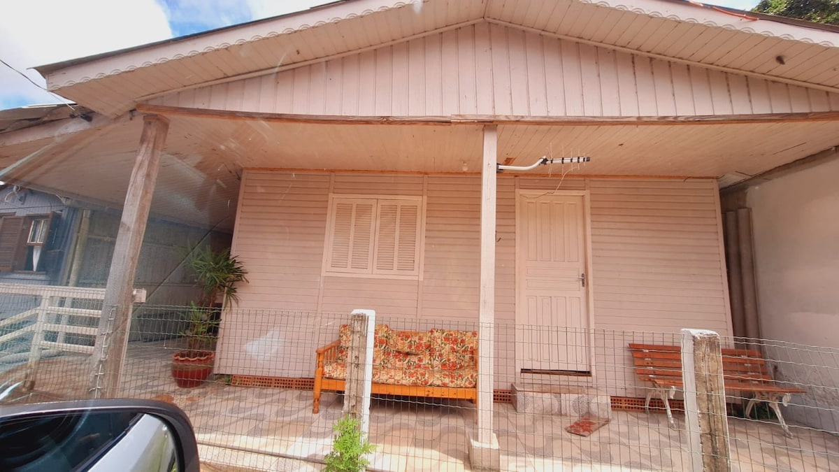 A single-story house with a light-colored wooden exterior is featured. The entrance is marked by a sturdy door and a large window, providing natural light. A decorative bench and a potted plant enhance the front porch area, bordered by a low fence.