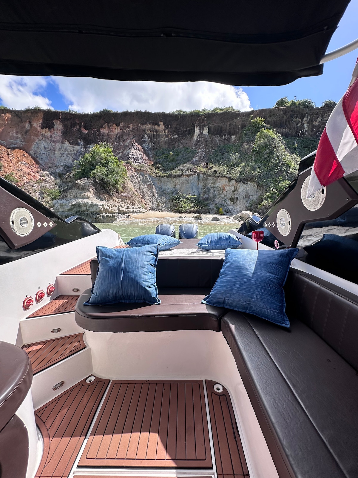 The interior of a boat features comfortable seating adorned with blue cushions. The dark wood flooring contrasts with the light-colored walls. In the background, natural cliffs rise dramatically, surrounded by greenery under a clear sky.