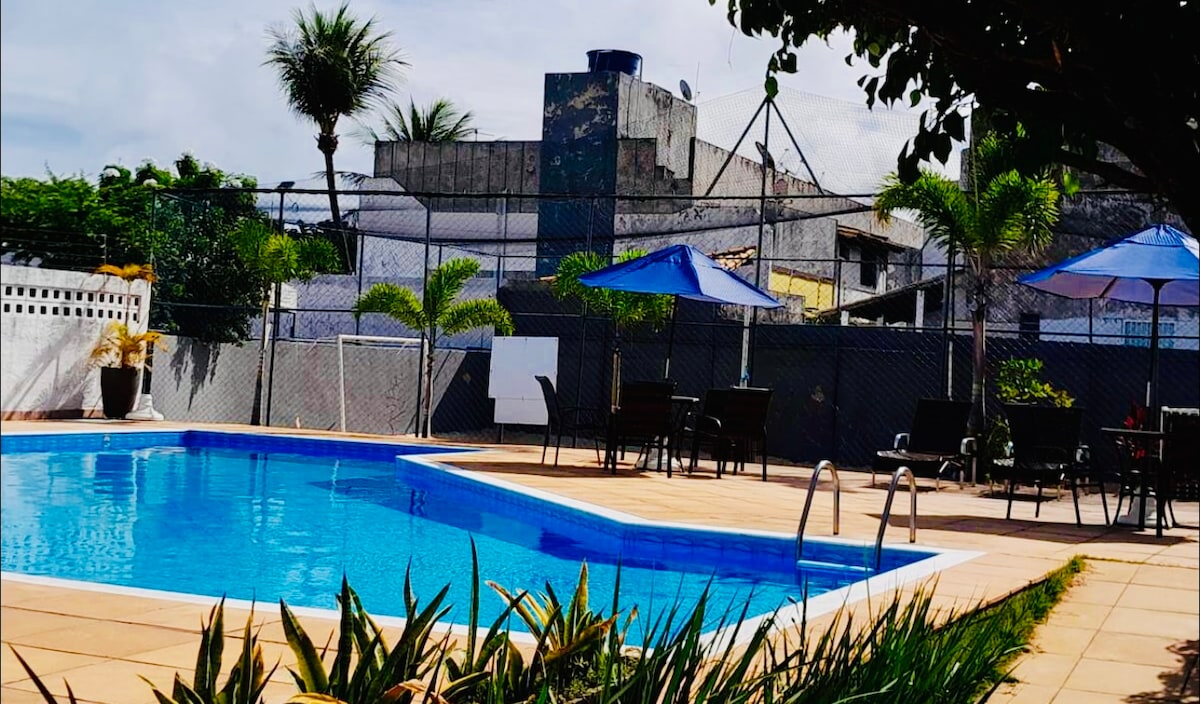 A large outdoor swimming pool is bordered by a tiled deck. Lounge chairs and umbrellas provide shade, and palm trees offer a tropical feel. A fence surrounds the area, ensuring privacy, while the surrounding buildings are partially visible in the background.