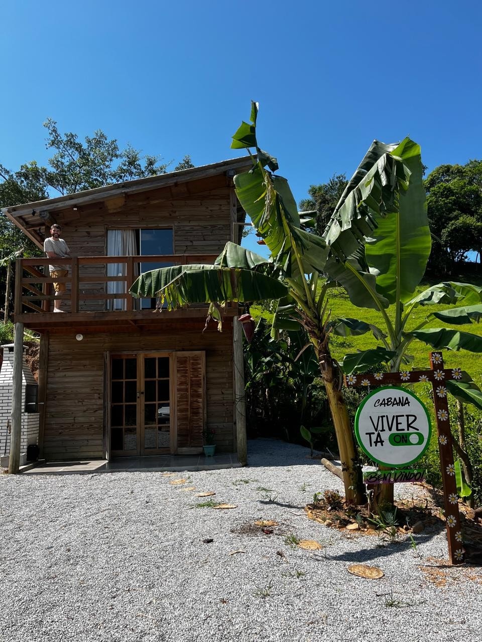 A rustic cabin made of wood is showcased under a clear blue sky. The structure features a second-floor balcony with sliding glass doors. Lush banana plants surround the entrance, and a sign indicating 'Cabana Viver' is visible, welcoming guests to this natural retreat.