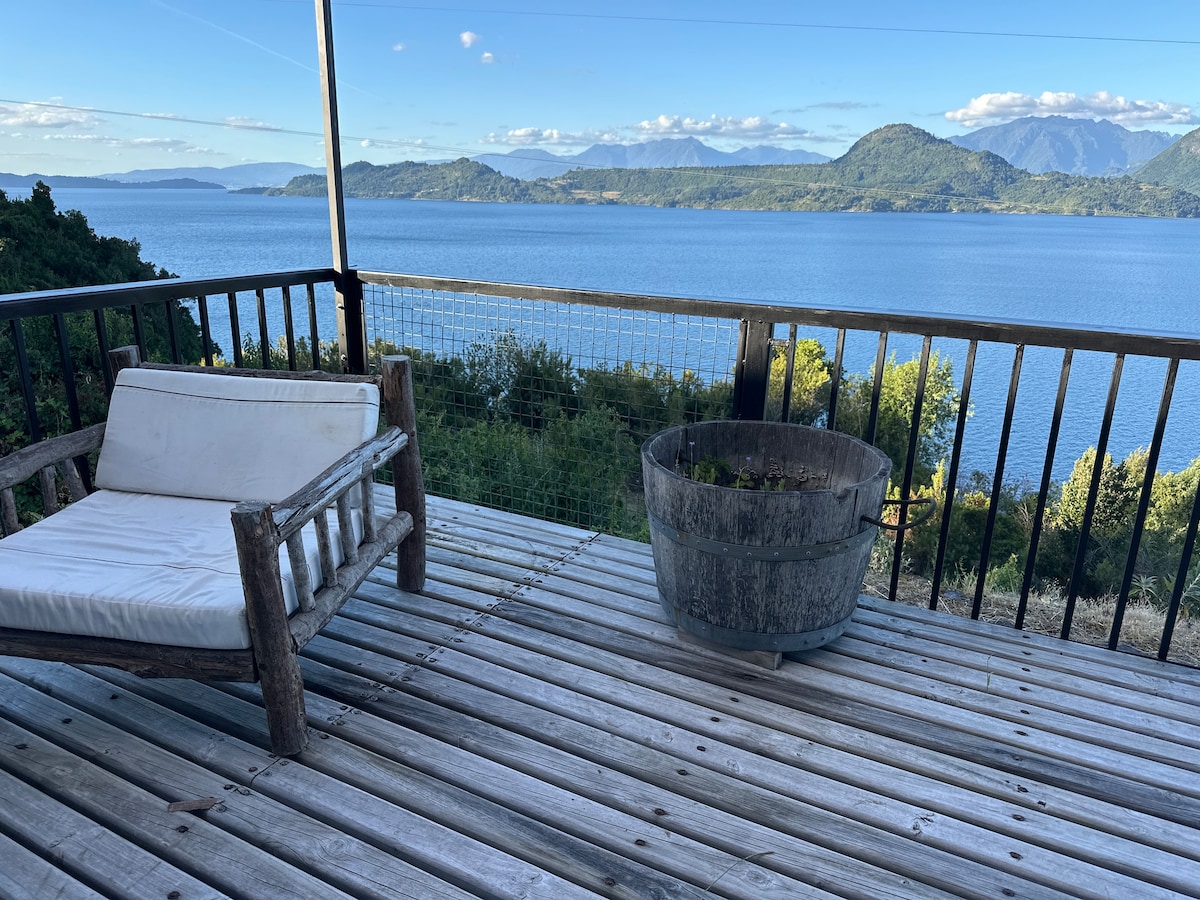 A spacious wooden terrace offers a view of Lago Ranco, complemented by surrounding greenery and distant mountains. A rustic wooden chair is positioned on the decking, alongside a circular planter made of metal. The serene water reflects the blue sky, enhancing the tranquil setting.