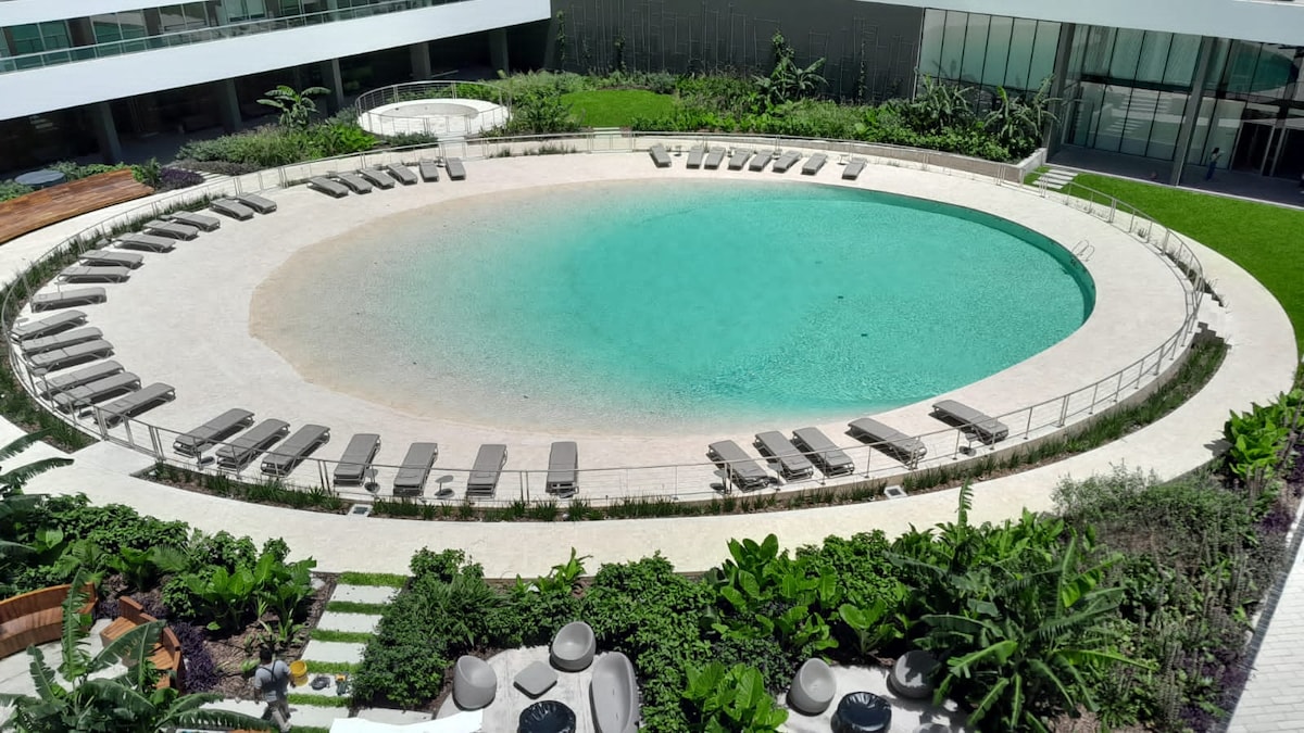An oval-shaped pool sits at the center of a green space, surrounded by numerous lounge chairs arranged in a semicircle. Lush plants and greenery are interspersed around the pool area, with smooth pathways leading towards the building in the background.