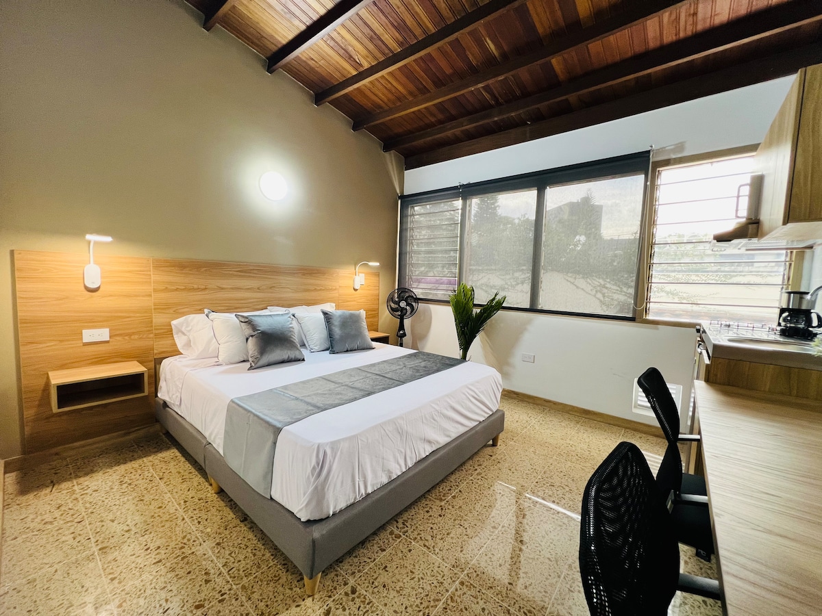 A spacious bedroom features a queen-sized bed dressed in neutral linens and accent pillows. Natural light fills the room through large windows, complemented by a ceiling fan and a potted plant near a kitchen area. Warm wooden beams enhance the inviting atmosphere.