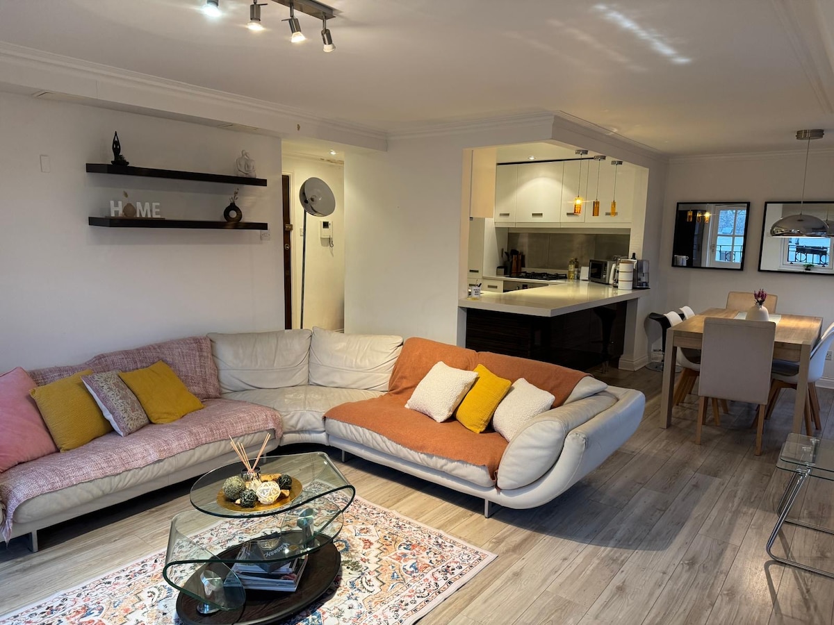 The living area features two modern sofas adorned with colorful cushions, positioned around a decorative coffee table. A dining table with chairs is visible in the background, complemented by an open kitchen area. Light fixtures enhance the bright, welcoming environment.