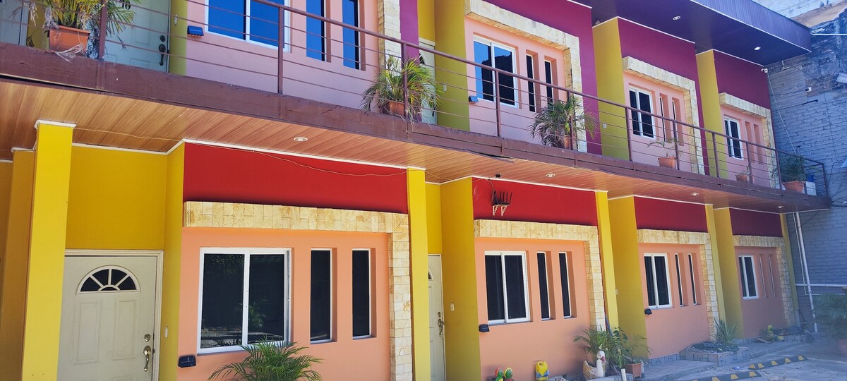 A multi-unit building features a vibrant exterior with a combination of orange, red, and yellow hues. Each unit is equipped with large windows and decorative planters. A welcoming entrance is visible, complemented by potted plants lining the walkway.