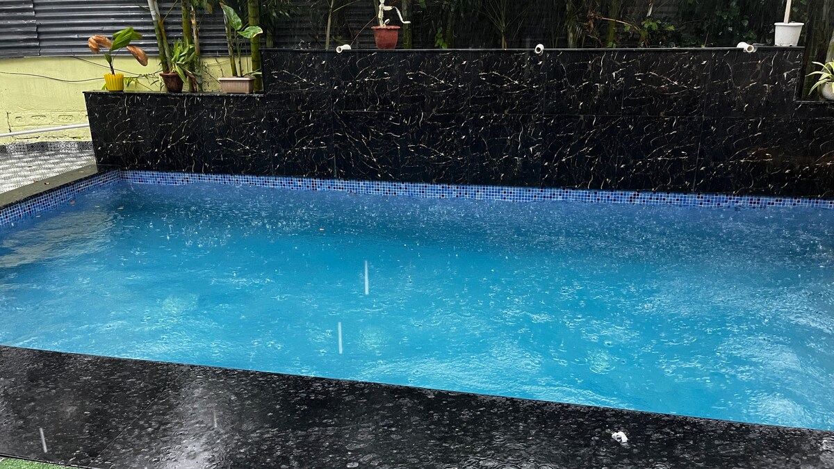 A swimming pool is seen with clear blue water, surrounded by a dark stone border. Rain falls gently on the surface, creating ripples. Lush greenery and potted plants are visible along the edges, adding a natural touch to the tranquil space.