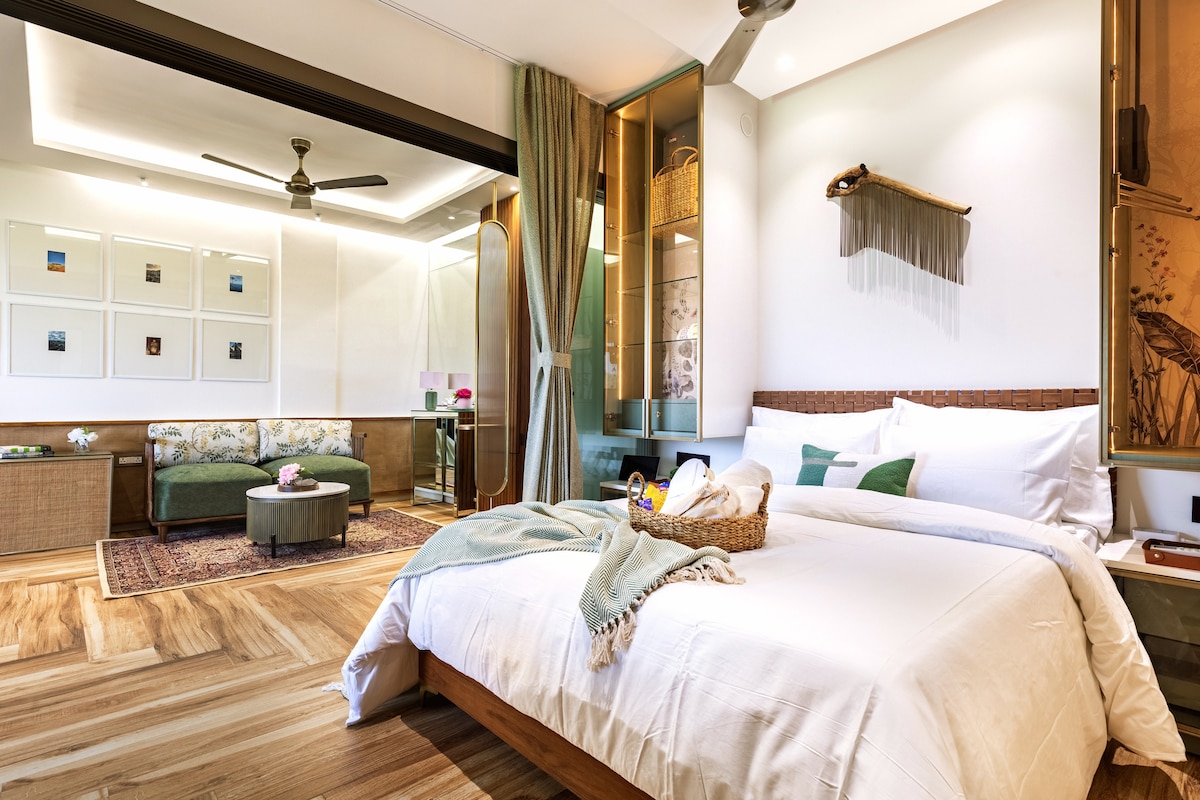 A spacious sleeping area showcases a large bed with fresh white linens and a green accent. In the background, a cozy seating area features a patterned sofa and a round coffee table, illuminated by soft lighting from the ceiling.