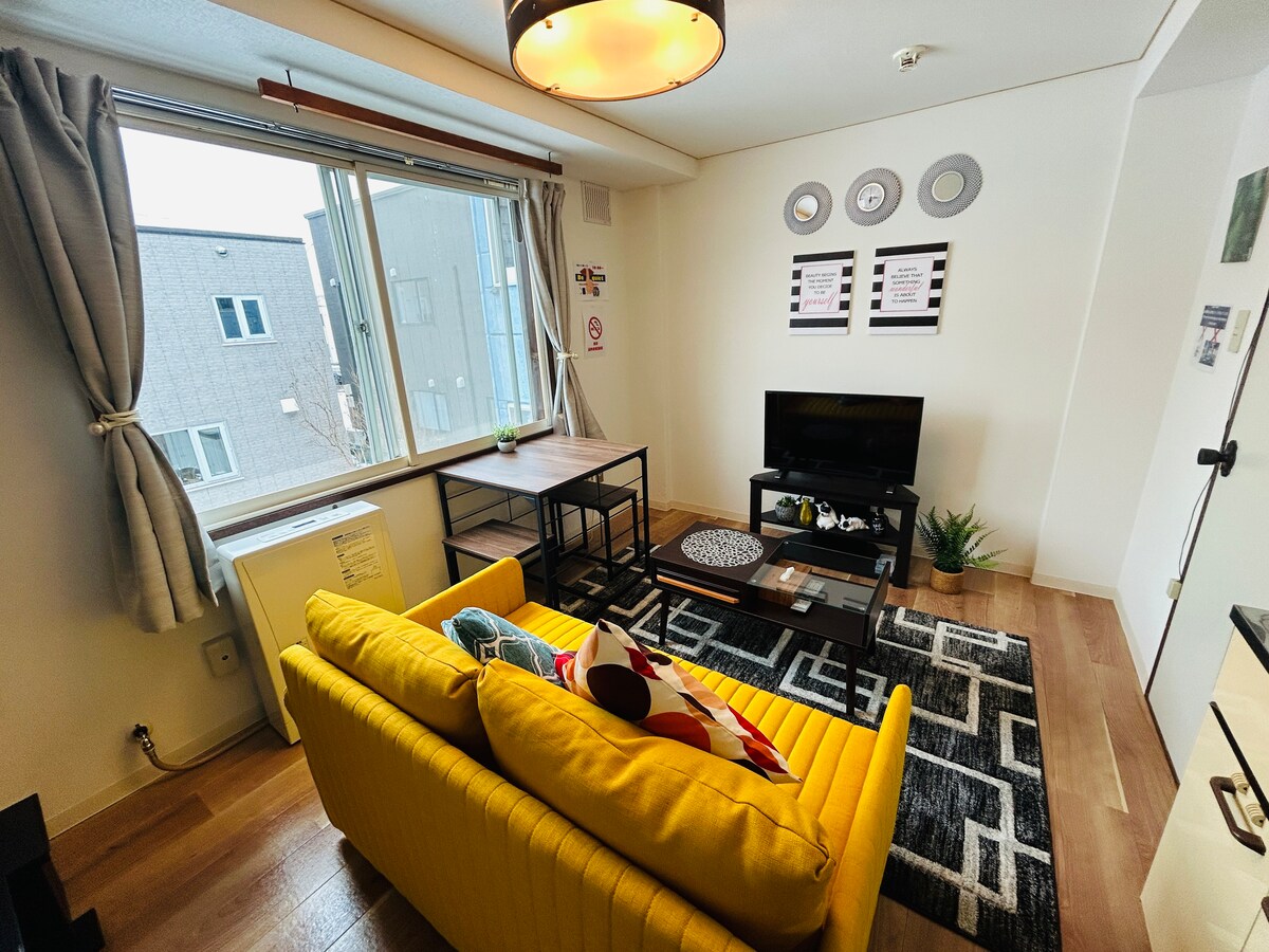 A bright living area features a yellow sofa, contrasted by a dark patterned rug. A wooden table and a TV stand hold a small television for entertainment. Natural light enters through a large window, enhancing the space's inviting design.