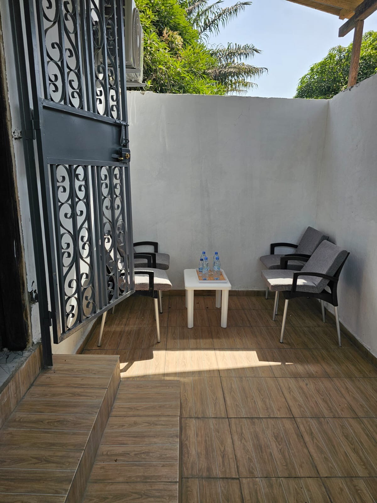A small terrace is viewed, featuring two armchairs and a table positioned on tiled flooring. Two bottles of water rest on the table. The surrounding walls are painted white, and greenery is visible above, suggesting a connection to the outdoors.