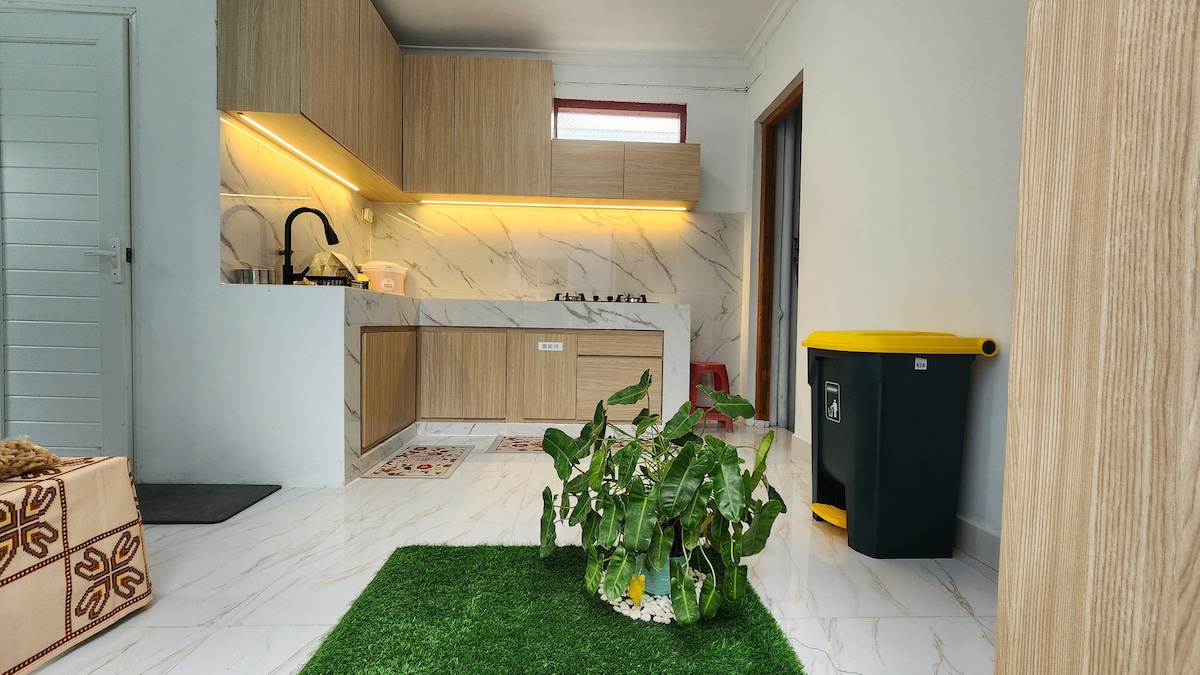 A modern kitchen area features wooden cabinetry complemented by a sleek countertop. Natural light enters through a window, illuminating the space. A green trash bin stands beside the cabinetry, while a small indoor garden with greenery adds a touch of nature to the room.