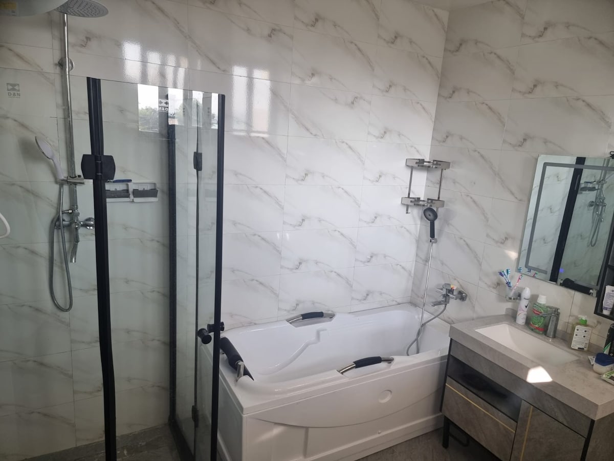 A modern bathroom features a glass-enclosed shower area alongside a stylish bathtub. Marble-patterned tiles cover the walls, and a sleek vanity with drawers is visible. A mirror overhead complements the clean lines and neutral color palette of the space, creating a functional bathing area.
