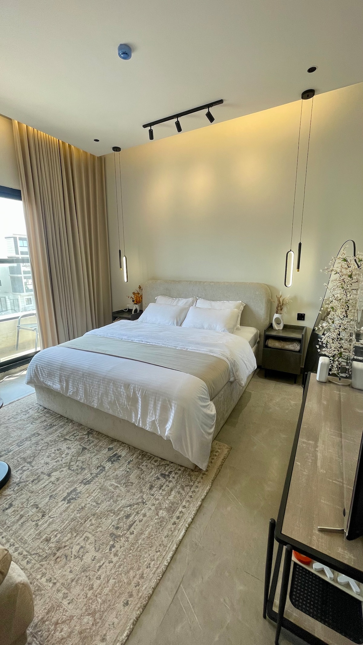 A bright room features a spacious bed with crisp white linens. Soft lighting is provided by modern pendant lamps hanging above. Floor-to-ceiling curtains frame the large window, while decorative elements on a nearby dresser add to the contemporary aesthetic.