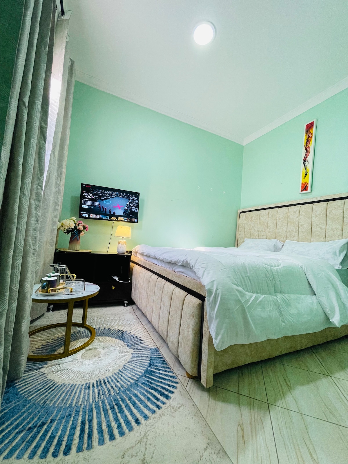 A clean and modern sleeping area features a spacious bed with crisp white linens. A small round table is positioned beside the bed, accompanied by a decorative rug. A Smart TV is mounted on the wall, providing convenience for guests. Soft natural light enters through the window, covered by light curtains.