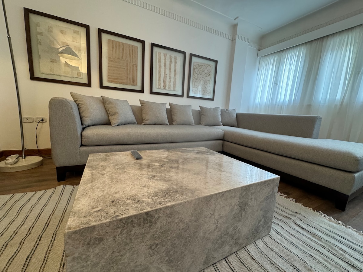 A modern living area features a spacious gray sectional sofa adorned with multiple cushions, arranged around a rectangular coffee table made of marble. Light-colored artwork is displayed on the walls, complemented by large windows dressed in sheer curtains. A textured area rug anchors the space.