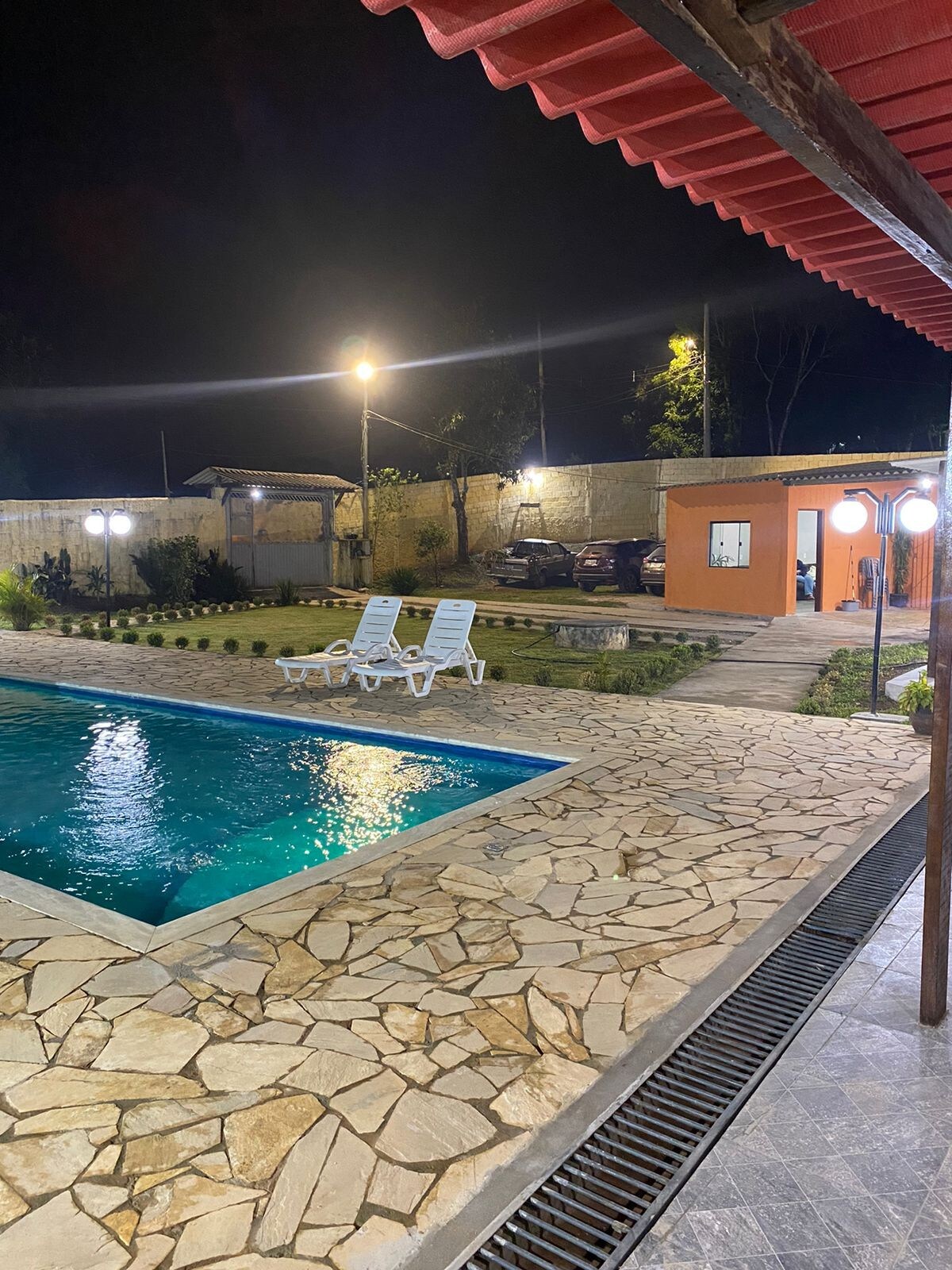 A nighttime view of a well-lit outdoor area featuring a swimming pool surrounded by a stone deck. Two lounge chairs are positioned near the pool, complemented by pathway lighting and landscaped greenery. A separate structure is visible in the background.
