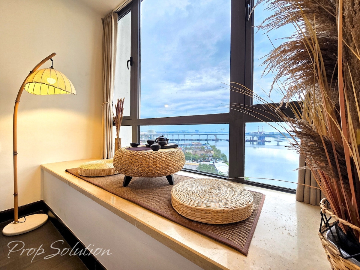 A cozy seating area is positioned by large windows, offering a clear view of the waterfront. Natural light enhances the space, which features a round table surrounded by woven seating cushions. A stylish floor lamp stands nearby, adding warmth to the modern decor.