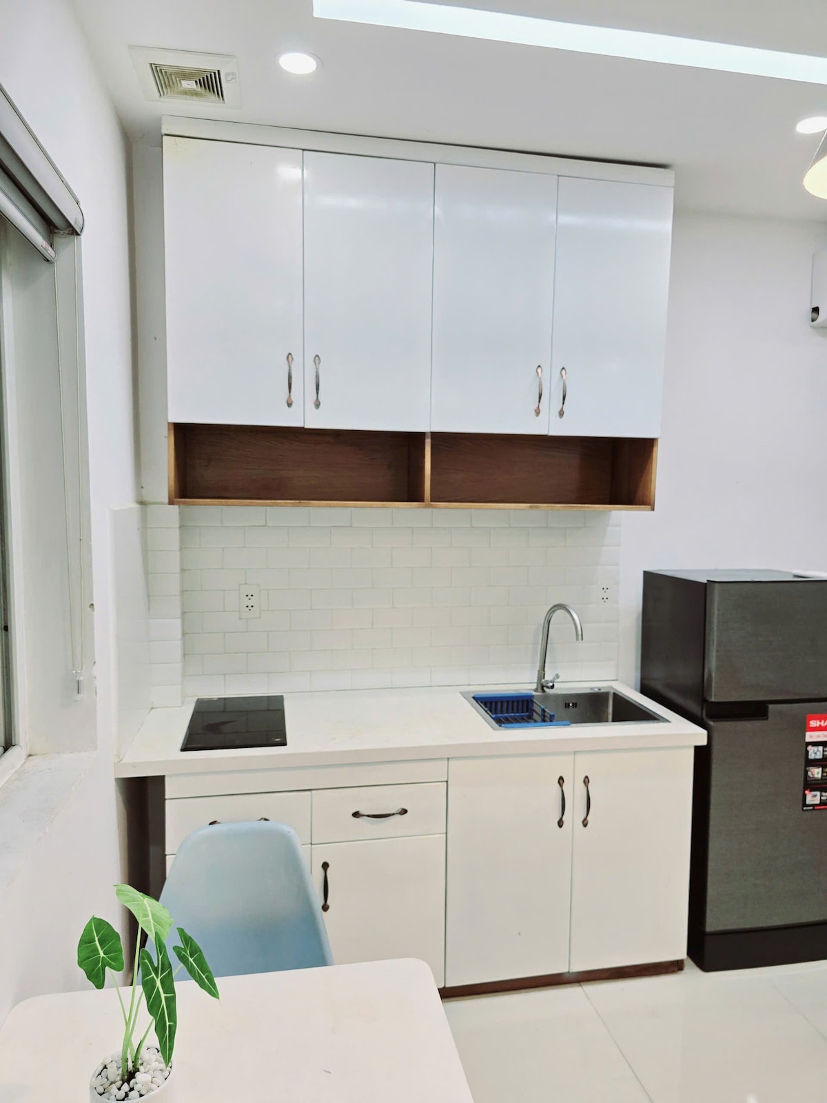 A modern kitchen area features sleek white cabinetry with a mix of upper and lower units. A stainless steel sink is set against white tiled walls, complemented by a built-in cooktop. A refrigerator stands to the right, completing the space. A small potted plant adds a touch of greenery.