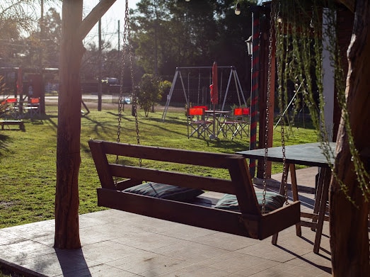 A wooden swing seat is suspended from a sturdy tree, featuring soft cushions for comfort. In the background, a grassy area hosts playground equipment and red picnic tables, providing an inviting space for relaxation and outdoor activities.