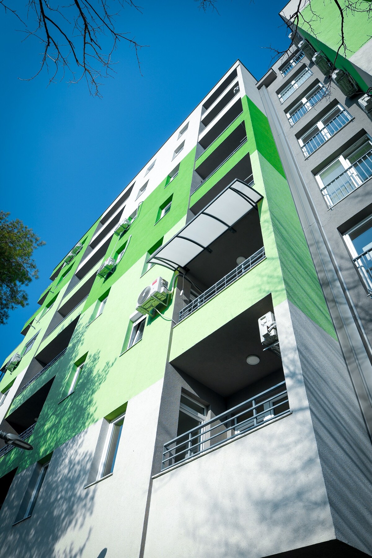A modern apartment building is depicted, featuring a unique green and gray façade. Balconies with railings are visible, along with several windows that reflect the clear blue sky. Foliage from nearby trees casts soft shadows on the exterior.