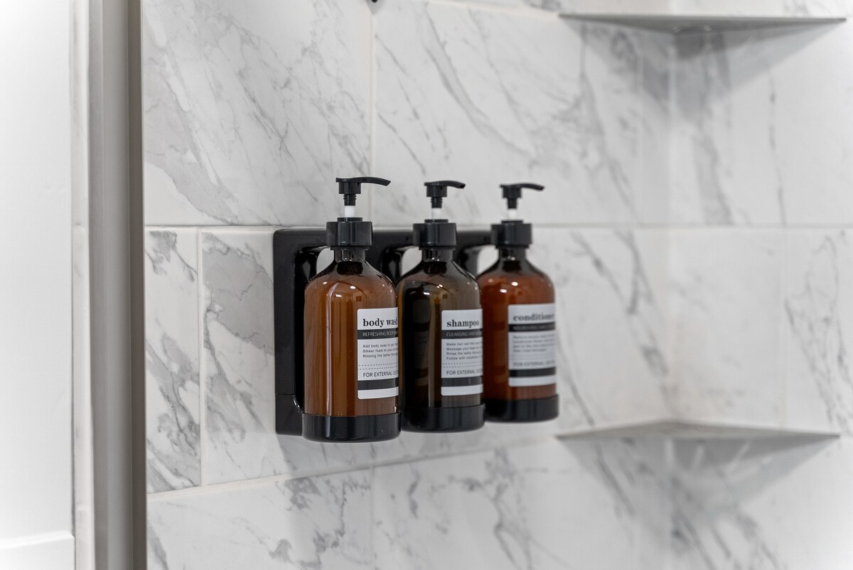 Three elegantly designed bottles are mounted on a marble wall, containing body wash, shampoo, and conditioner. Shelves are visible in the background, providing additional storage for toiletries.