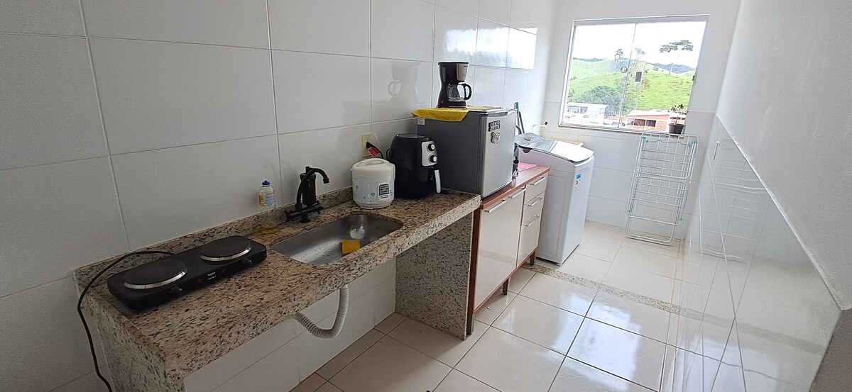 A modern kitchen features a white tiled wall and granite countertops. Essential appliances, including a coffee maker and washing machine, are arranged neatly. A window offers a view of greenery outside, enhancing natural light in the space.