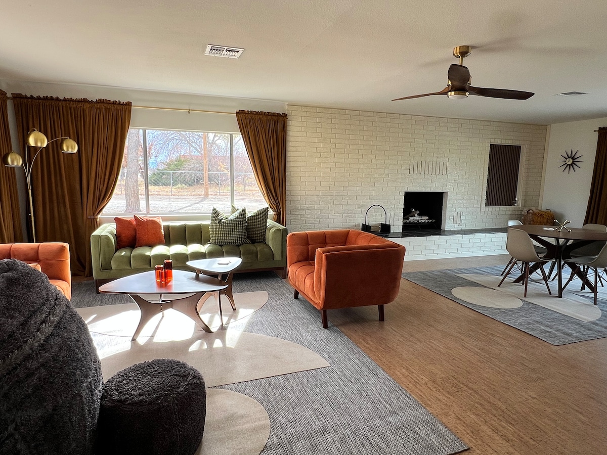 A spacious living area is highlighted by large windows that let in natural light, showcasing the mid-century furnishings. Cozy seating is provided by a green sofa and two orange armchairs, arranged around a modern coffee table. A dining area with stylish chairs sits adjacent to the seating area.