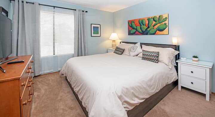 This quiet corner bedroom offers a king-sized bed, ceiling fan, smart TV, charging ports, 2 nightstands, a dresser, and a closet. There are also two large windows with blackout curtains.