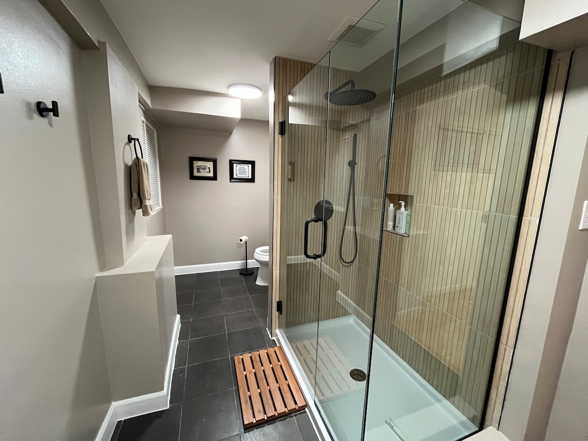 A modern bathroom features a spacious shower with a glass enclosure and wooden bath mat. Soft lighting brightens the neutral-toned walls and floor tiles. A framed picture hangs above a small toilet, while a complementary towel is neatly hung on a nearby hook.