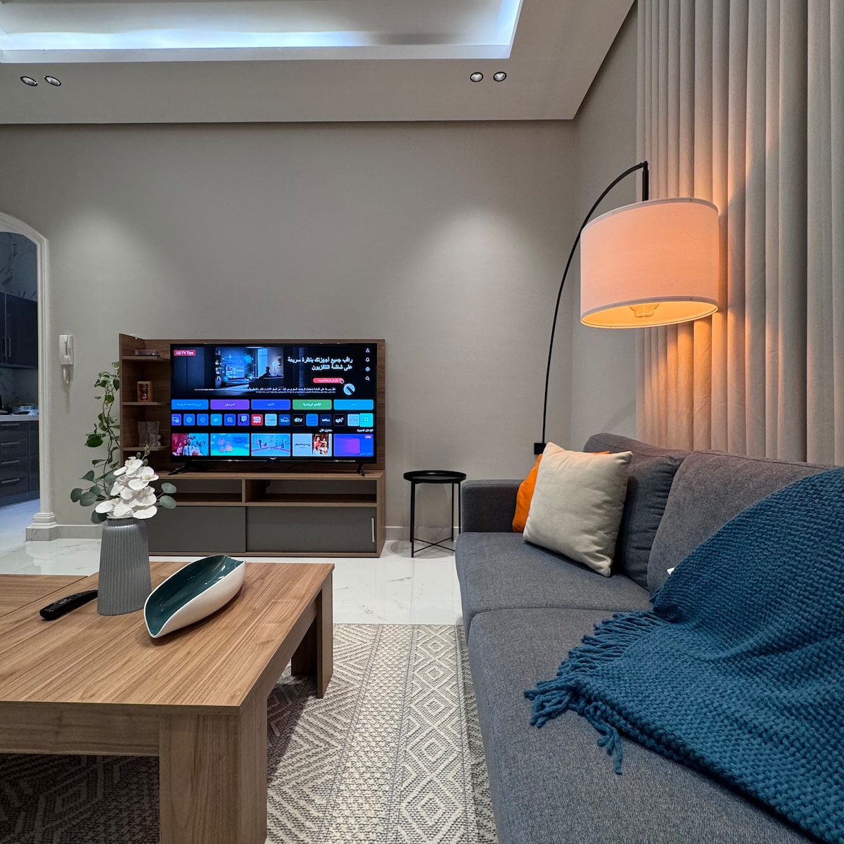 A modern living area is presented with a soft gray sofa adorned with textured pillows and a cozy throw. A wooden coffee table sits in front, accompanied by an elegant decorative vase. A wall-mounted TV is visible alongside a stylish lamp, casting warm light on the space.