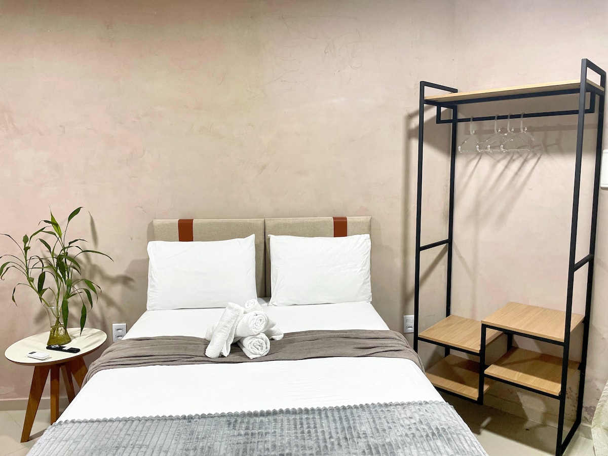 A neatly arranged sleeping area features a spacious bed with white linens and decorative gray accents. Next to the bed, a small table holds a potted plant. A sturdy clothing rack is visible, showcasing hanging hooks and a wooden shelf.