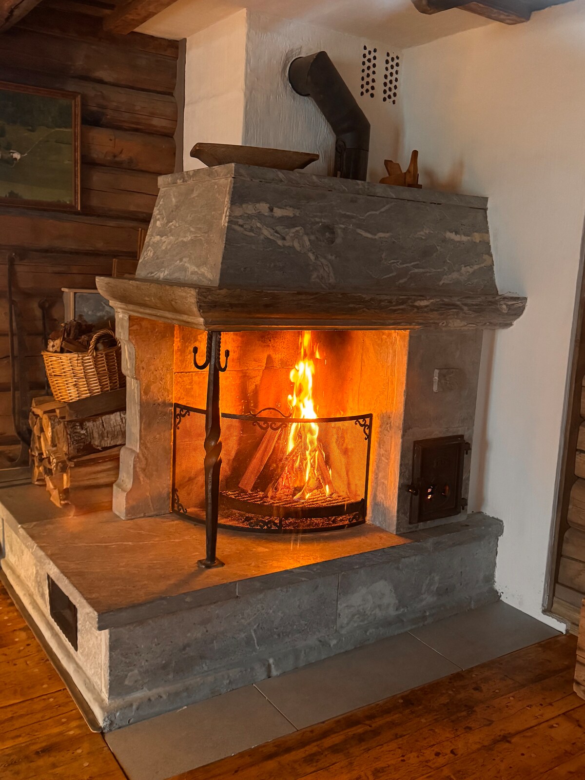 A stone fireplace is featured, emanating a warm glow from the flames within. The fireplace is framed by a rustic wood interior, complementing the cozy atmosphere. Nearby, a basket is positioned, suggesting a supply of firewood for comfort and warmth during cooler evenings.
