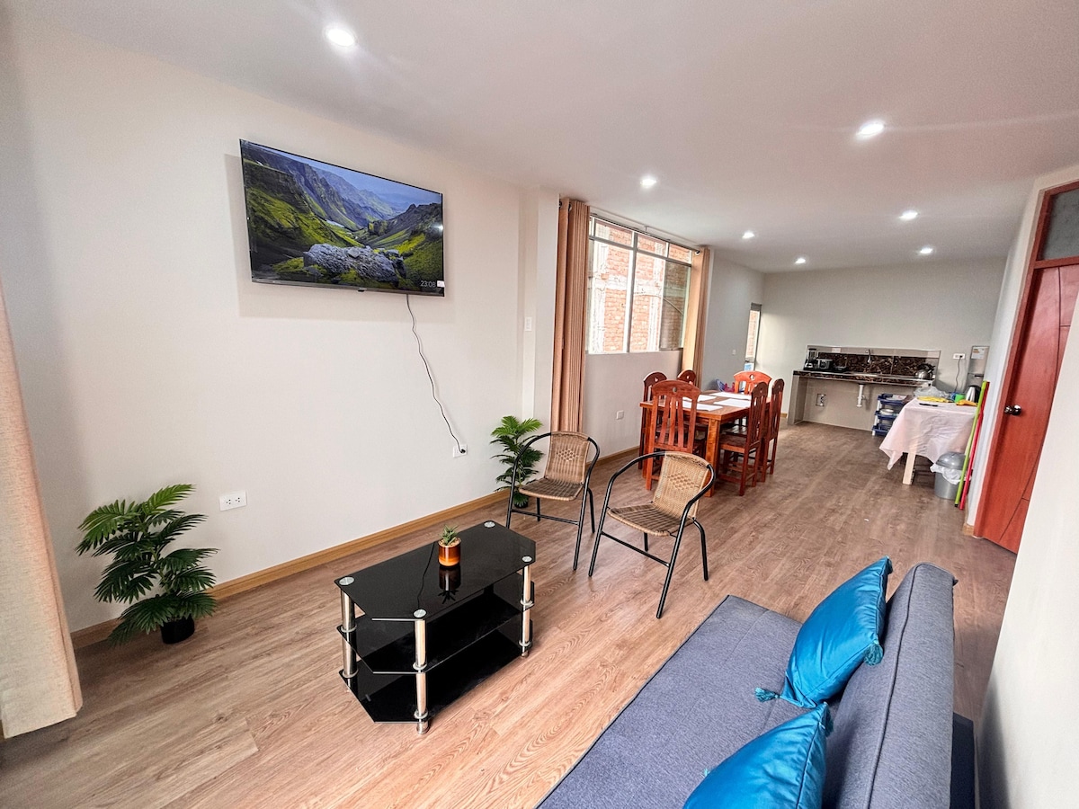 A welcoming living area features a comfortable sofa with blue cushions facing a glass coffee table. A flat-screen TV is mounted on the wall, while a dining table with chairs is located in the background, adjacent to a well-equipped kitchen with visible cabinetry.