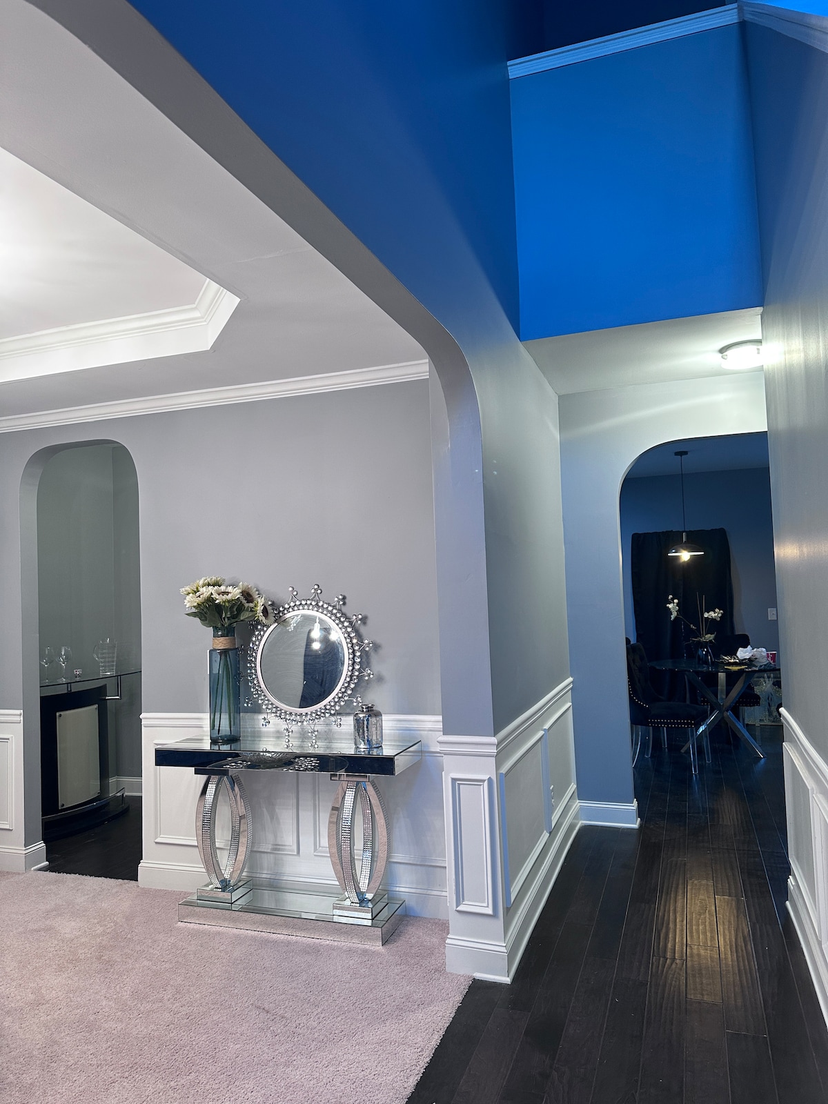 A stylish entryway features light grey walls contrasted by a vibrant blue ceiling. A round mirror rests on a sleek console table, accompanied by a vase of flowers. To the left, an archway leads to a room, while a hallway extends to the right.