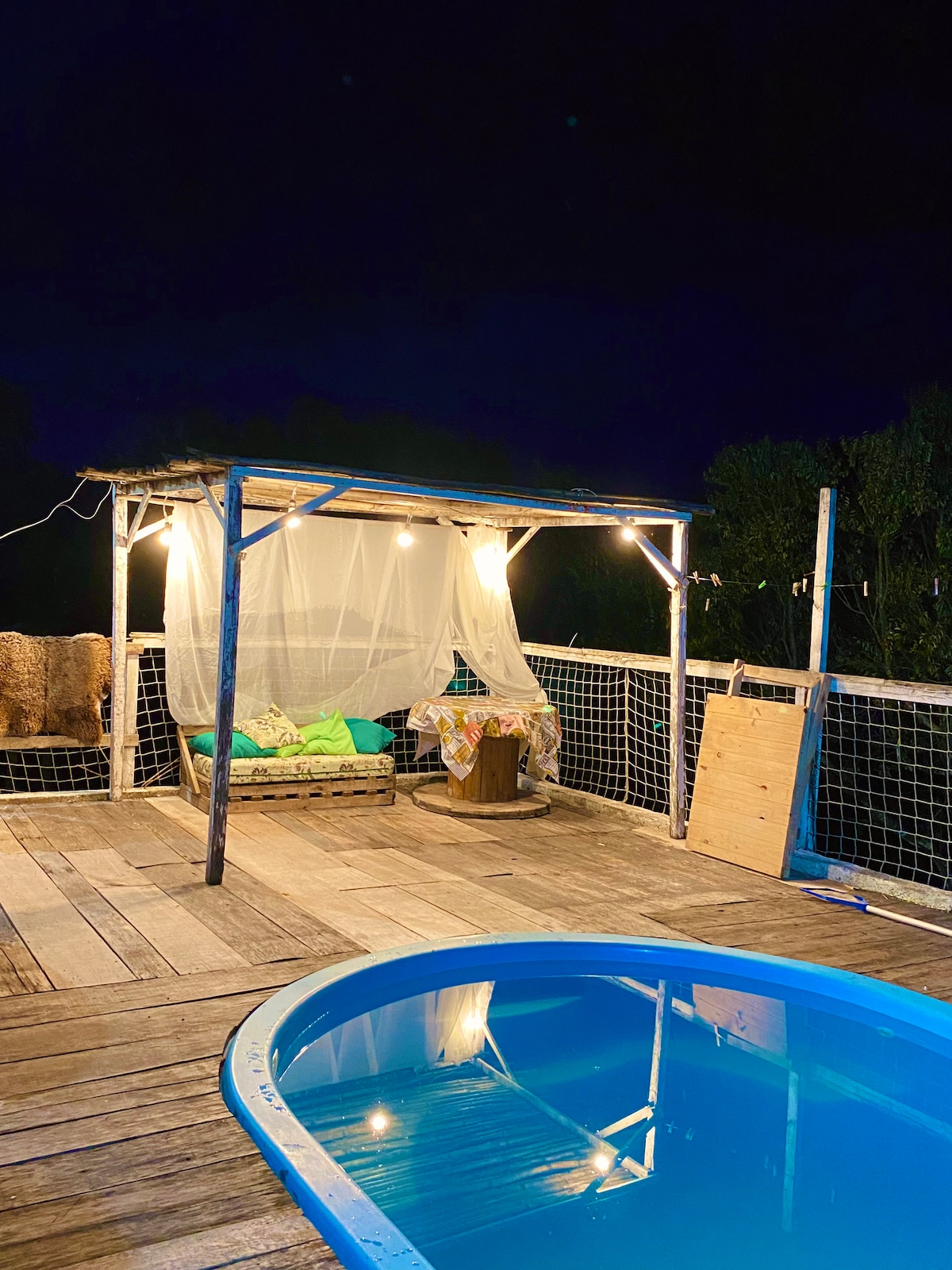 An outdoor space features a small, circular pool beside a wooden deck. A cozy seating area, partially draped in a light fabric, provides a tranquil spot for relaxation. Soft lighting creates an inviting ambiance against the night sky, with lush greenery surrounding the area.