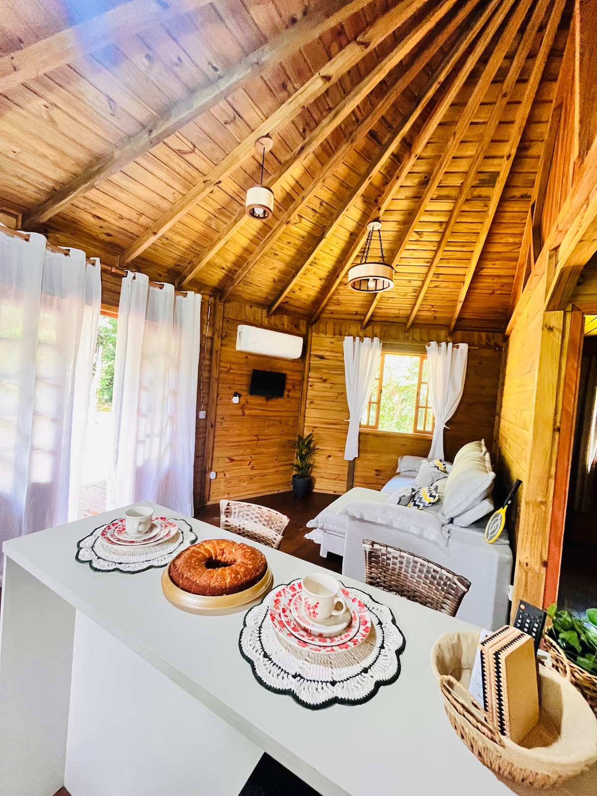 The interior of the cabana features a wooden ceiling with a rustic charm. A white table is set with decorative placemats and a cake on display. A cozy seating area includes a light-colored sofa, complemented by natural light from the windows, enhancing the warm atmosphere.