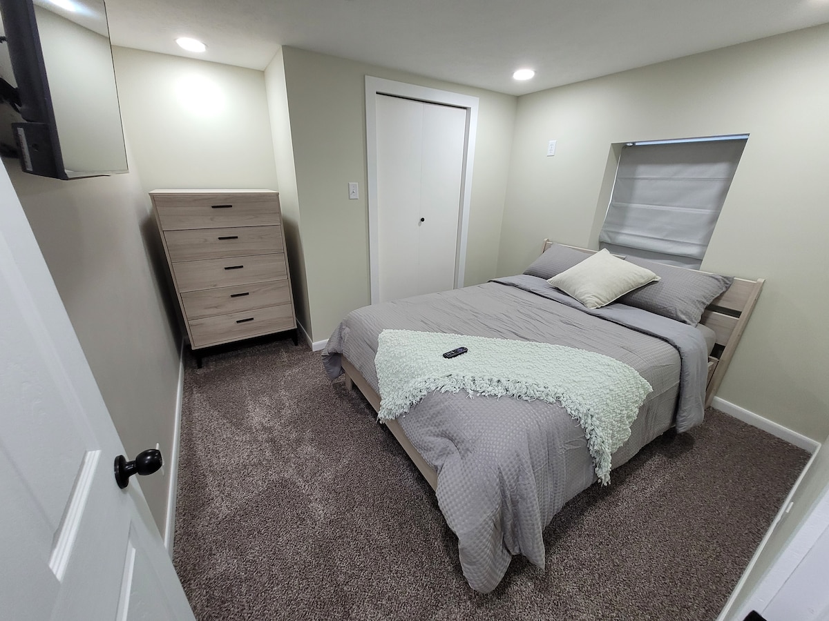 Сутеренна спалня №2 Изходен прозорец, пълноразмерно легло, телевизор с дистанционно управление, скрин и килер със закачалки.