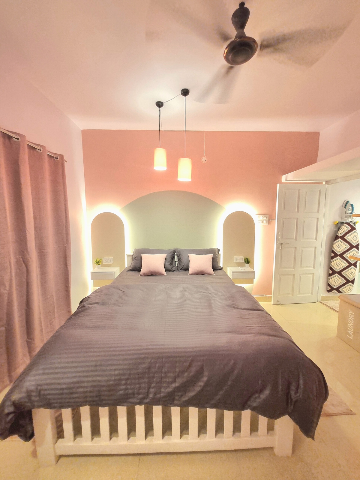 A cozy bedroom features a spacious bed with dark bedding and decorative pillows. Soft lighting is provided by two hanging lamps above the headboard, and a fan is mounted on the ceiling. Two nightstands are situated on either side of the bed, and the room includes light pink and green accent walls.