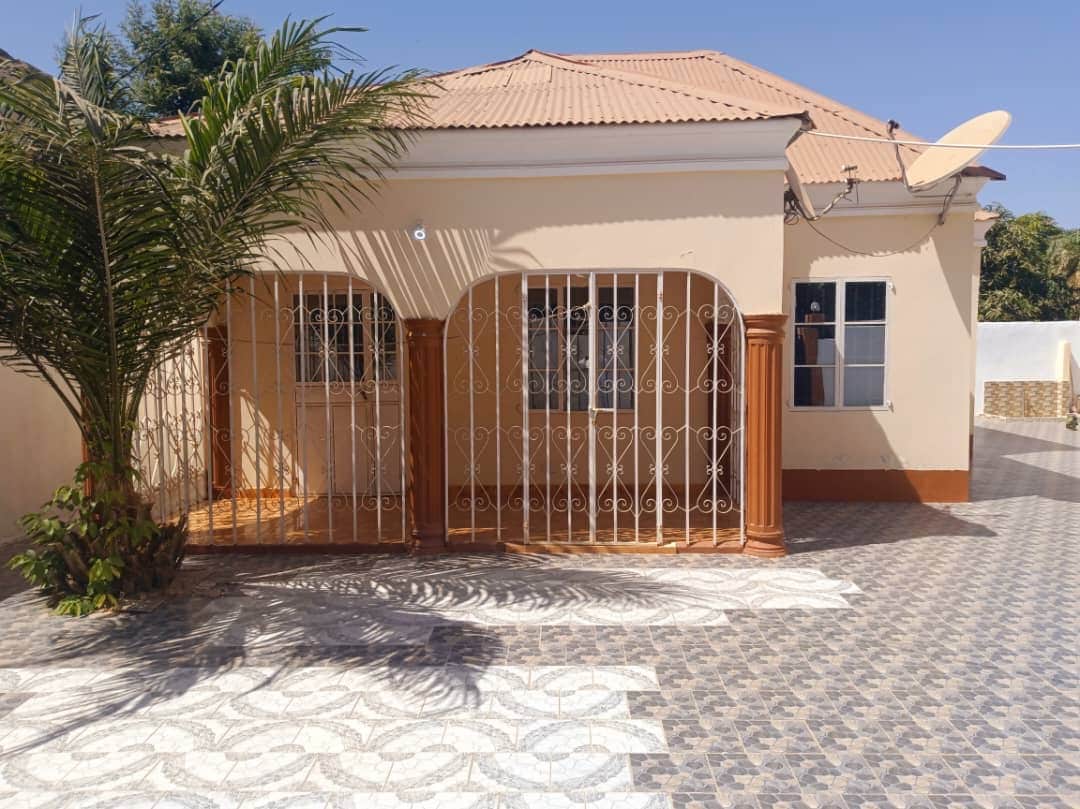 A single-story building features a front porch enclosed by decorative metal bars. Large windows allow natural light to fill the living space. A patterned tiled pathway leads to the entrance, which is flanked by greenery and a palm tree.