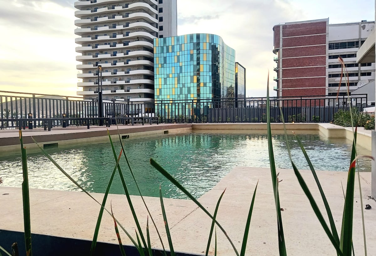 A sleek, modern pool is bordered by a clean stone deck, surrounded by greenery. Tall buildings with reflective glass facades are visible in the background, showcasing a blend of urban architecture against the setting sky.